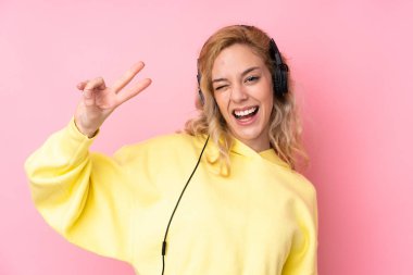 Pembe arka planda izole edilmiş sweatshirt giyen genç sarışın kadın müzik dinliyor ve şarkı söylüyor.