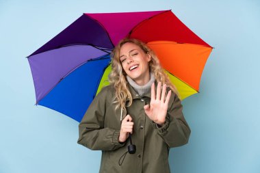 Mavi arka planda şemsiye tutan genç sarışın kadın mutlu bir ifadeyle selamlıyor.