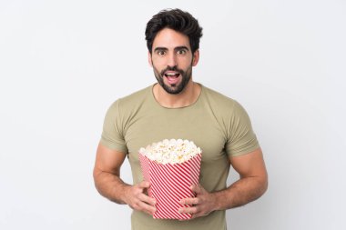 Soyutlanmış beyaz arka planda sakallı yakışıklı genç bir adam elinde koca bir kova patlamış mısır tutuyor.