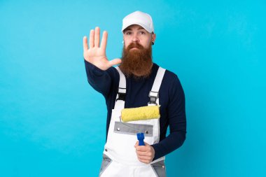 Uzun sakallı bir ressam. Mavi arka planda. Elleriyle jesti durduruyor.