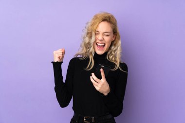 Mor arka planda izole edilmiş genç sarışın kadın, telefon zafer pozisyonunda.