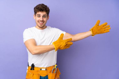 Mor arka planda izole edilmiş genç elektrikçi, davet edilmek için ellerini yana uzatıyor.