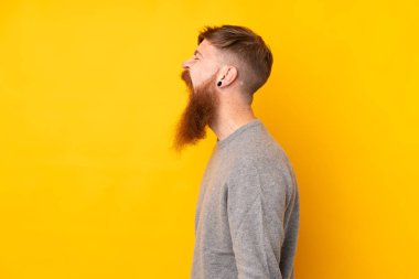 Sarı arka planda uzun sakallı kızıl saçlı adam yan pozisyonda gülüyor.