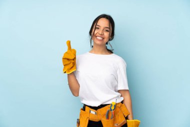 Genç elektrikçi kadın mavi arka planda izole edilmiş ve parmağını kaldırıyor.