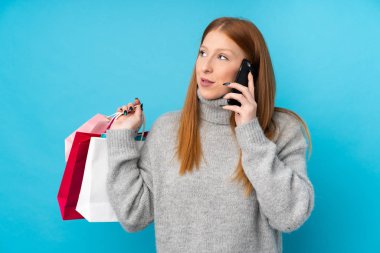 İzole edilmiş mavi arka planda genç kızıl saçlı kadın elinde alışveriş çantaları ve cep telefonuyla arkadaşını arıyor.