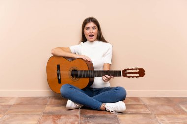 Gitarlı genç bir kadın yerde oturuyor ve yüzünde sürpriz bir ifade var.