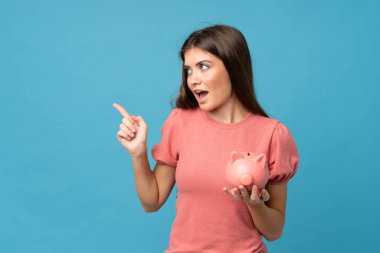 Soyutlanmış mavi arka planı olan genç bir kadın büyük bir kumbara tutuyor.