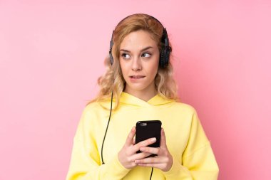 Pembe arka planda izole edilmiş sweatshirt giyen genç sarışın kadın cep telefonuyla müzik dinliyor ve düşünüyor.