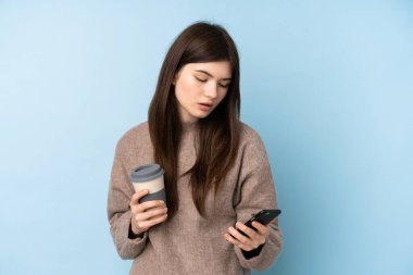 İzole edilmiş mavi arka plan üzerine kazak giyen genç Ukraynalı kız elinde kahve ve bir cep telefonu tutuyor.