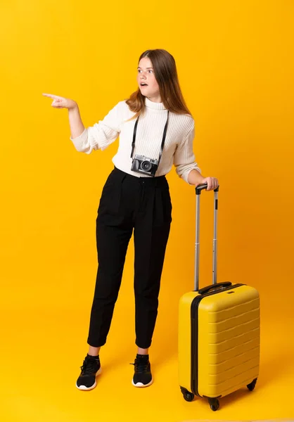 Full body of traveler teenager girl with suitcase over isolated yellow background pointing finger to the side