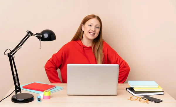 Young student woman in a workplace with a laptop posing with arms at hip and smiling