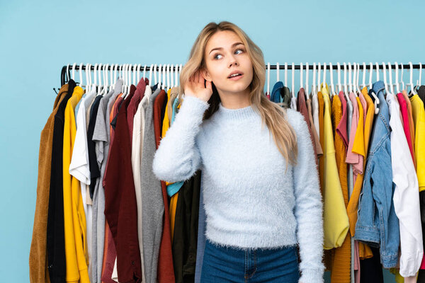 Teenager Russian girl buying some clothes isolated on blue background listening to something by putting hand on the ear