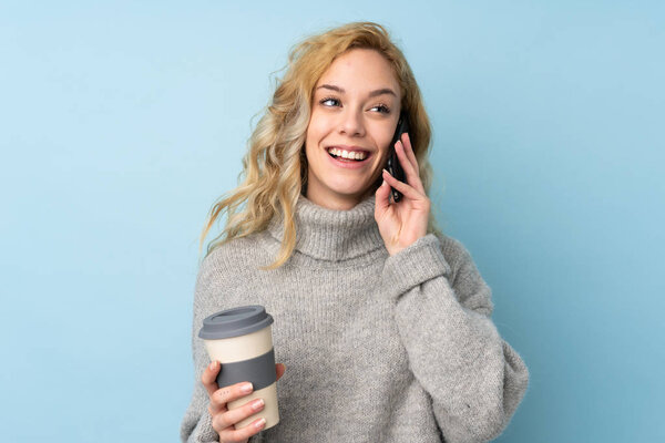 Young blonde woman wearing a sweater isolated on blue background holding coffee to take away and a mobile