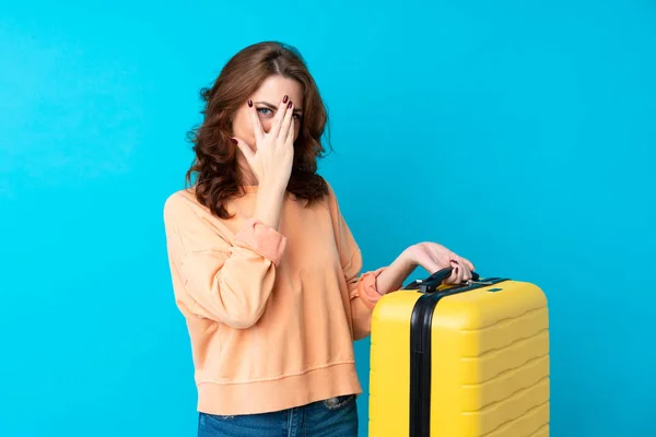 Traveler woman with suitcase over isolated blue background covering eyes and looking through fingers