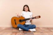 Mladá žena s kytarou sedící na podlaze a ukazující na laterály, která pochybuje