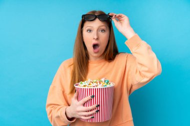 Genç kızıl saçlı, soyutlanmış arka planda 3D gözlüklü ve elinde koca bir kova patlamış mısır tutan bir kadın.