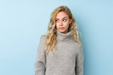 Mavi arka planda izole edilmiş süveter giyen genç sarışın kadın şüpheli hareketler yapıyor.