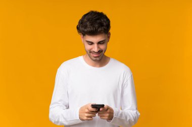 İzole edilmiş turuncu arka planı olan genç bir adam cep telefonuyla mesaj gönderiyor.