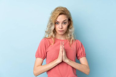 Mavi arka planda izole edilmiş genç sarışın kadın yalvarıyor.