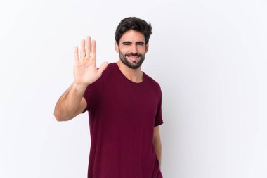 Sakallı yakışıklı genç adam izole edilmiş beyaz arka planda mutlu bir ifadeyle selam veriyor.