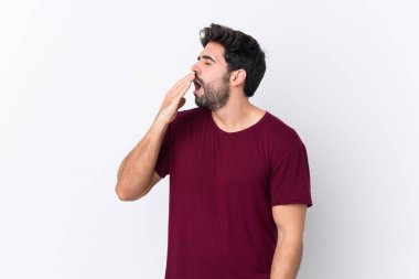 Sakallı yakışıklı genç bir adam izole edilmiş beyaz arka planda esniyor ve ağzını eliyle açık tutuyor.