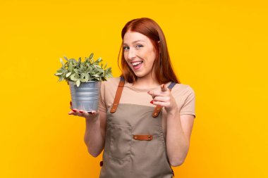 Genç, kızıl saçlı bahçıvan kadın, sarı arka plan üzerinde bir bitki tutuyor ve parmağını sana gösteriyor.