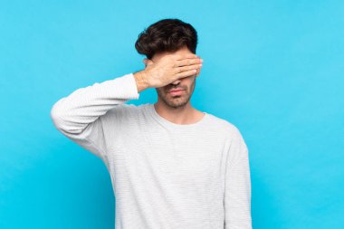 İzole edilmiş mavi arka planı olan genç bir adam gözleri elleriyle kapatıyor. Bir şey görmek istemiyorum.