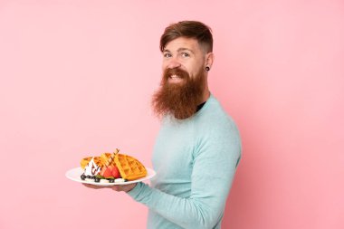 Kızıl saçlı, uzun sakallı bir adam izole edilmiş pembe arka planda waffle tutuyor ve gülümsüyor.
