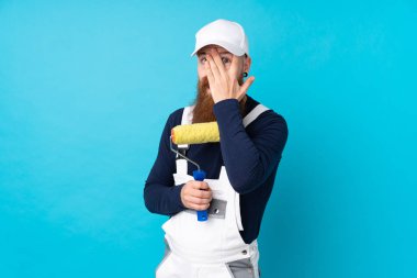 Uzun sakallı ressam, izole edilmiş mavi arka planda, gözleri kapatıyor ve parmaklarından bakıyor.