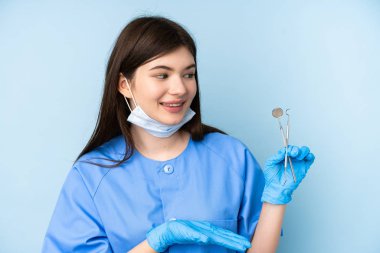 İzole edilmiş mavi arka planda aletleri tutan genç dişçi kadın davet etmek için ellerini yana uzatıyor.