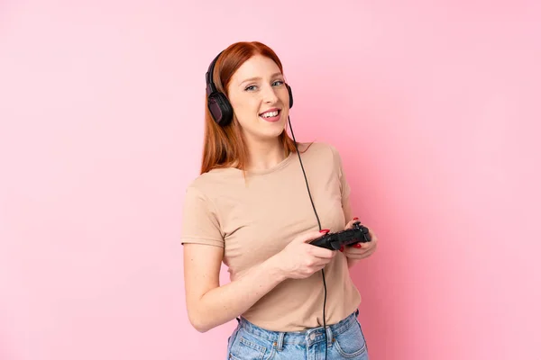 年轻的红头发女人在孤立的粉色背景上玩电子游戏 — 图库照片