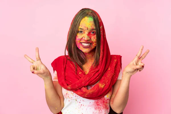 Young Indian woman with colorful holi powders on her face isolated on pink background showing victory sign with both hands