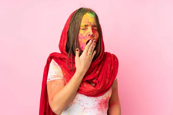 Young Indian woman with colorful holi powders on her face isolated on pink background yawning and covering wide open mouth with hand