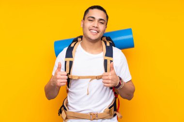 Sarı arka planda izole edilmiş büyük sırt çantalı Asyalı genç bir adam baş parmak işareti yapıyor.