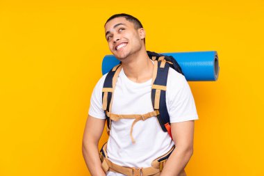 Sarı arka planda izole edilmiş büyük sırt çantalı Asyalı genç adam gülüyor ve yukarı bakıyor.