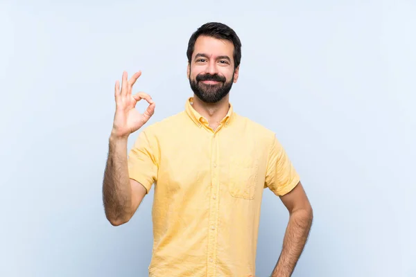 一个留着胡子的年轻人用手指在孤立的蓝色背景上表现出一个好的标志 — 图库照片