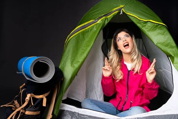 Teenager girl inside a camping green tent isolated on black background surprised and pointing up