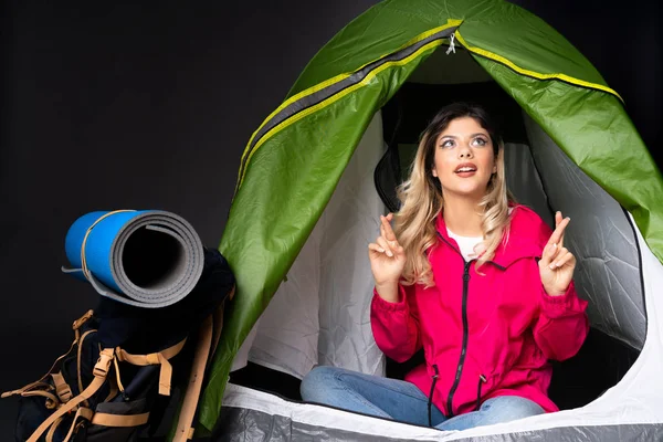 Teenager girl inside a camping green tent isolated on black background with fingers crossing and wishing the best