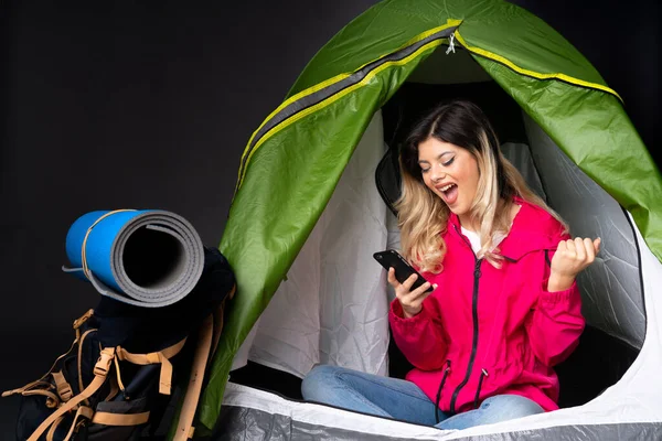Teenager girl inside a camping green tent isolated on black background surprised and sending a message