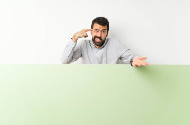 Sakallı, kocaman boş bir afiş tutan genç yakışıklı adam deliliğin jestini yapıyor. Parmağını kafaya koyuyor.