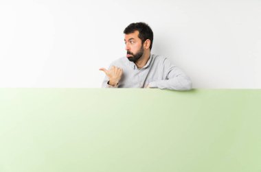 Sakallı genç yakışıklı adam elinde kocaman boş bir pankart tutuyor mutsuz ve bir tarafı işaret ediyor.