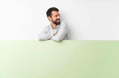 Sakallı genç yakışıklı adam kolları çapraz ve mutlu yeşil boş bir levha tutuyor.