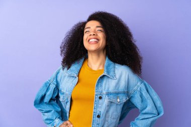 Genç Afrikalı Amerikalı bir kadın, mor arka planda izole edilmiş, kolları kalçasında ve gülümsüyor.