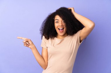 Mor arka planda izole edilmiş genç Afro-Amerikalı kadın şaşırmış ve işaret ediyor.