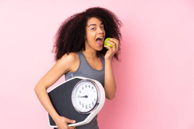Genç Afrikalı Amerikalı kadın pembe arka planda, tartı makinesi ve elma ile izole edilmiş.