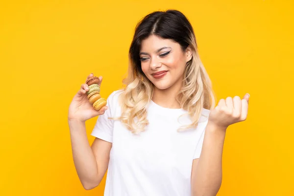 Menina Adolescente Isolado Fundo Amarelo Segurando Macarons Franceses Coloridos Apontando — Fotografia de Stock