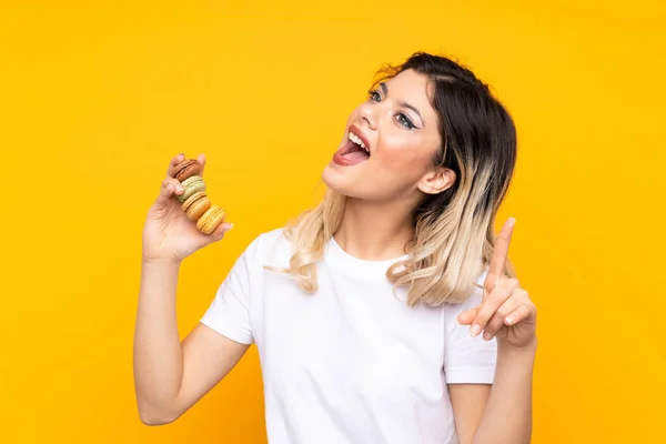 Menina Adolescente Isolada Fundo Amarelo Segurando Macarons Franceses Coloridos Apontando — Fotografia de Stock