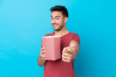 Soyutlanmış mavi arka planda genç yakışıklı bir adam elinde koca bir kova patlamış mısırla ön tarafı işaret ediyor.