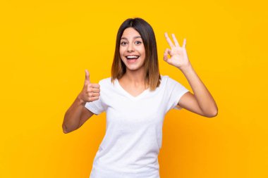 İzole edilmiş sarı arka plan üzerinde genç bir kadın tamam işareti ve baş parmak işareti gösteriyor.