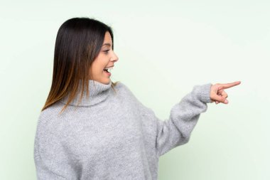 İzole edilmiş yeşil arka planda süveter giyen bir kadın parmağını yana doğru gösteriyor.